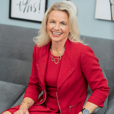 Smiling woman with blonde hair in red dress and jacket sitting on gray couch against pale blue wall.