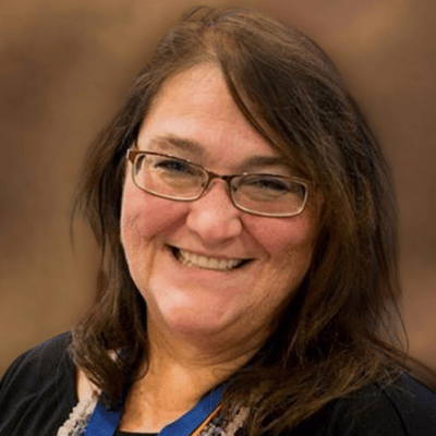 Smiling woman with glasses and long brown hair against a solid brown background.