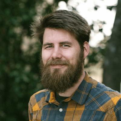 Bearded man with brown hair wearing a blue and yellow plaid shirt against a blurred outdoor background.