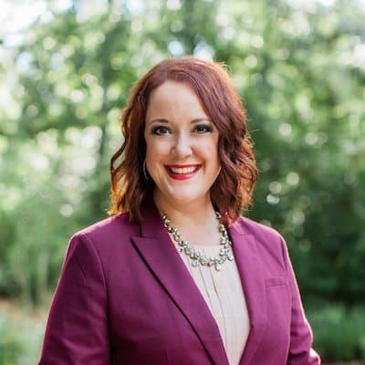 Smiling woman with curly red hair in a purple blazer and statement necklace outdoors with trees behind.