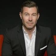 Man in a black suit jacket and white shirt sitting against a dark background.