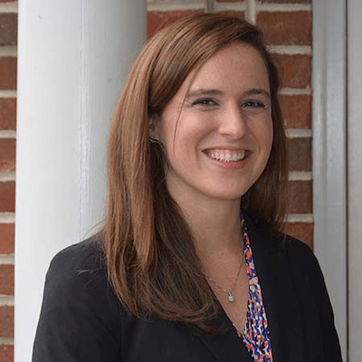 Smiling woman with long light brown hair wearing a black blazer and patterned blouse against a brick wall.
