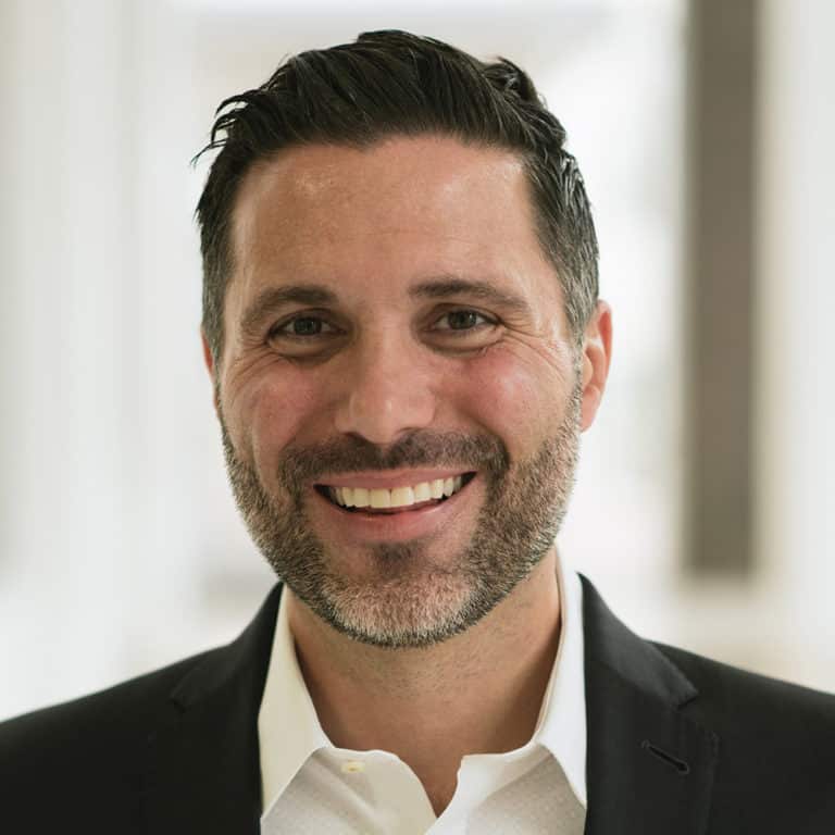 Smiling man with dark hair and beard wearing a black blazer and white shirt.