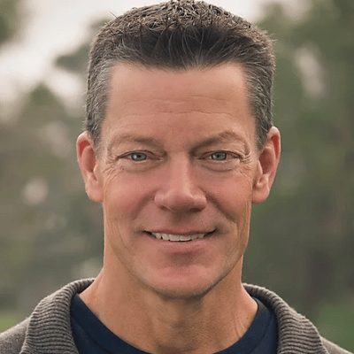 Smiling man with short dark hair wearing a brown sweater and blue shirt, blurred greenery background.