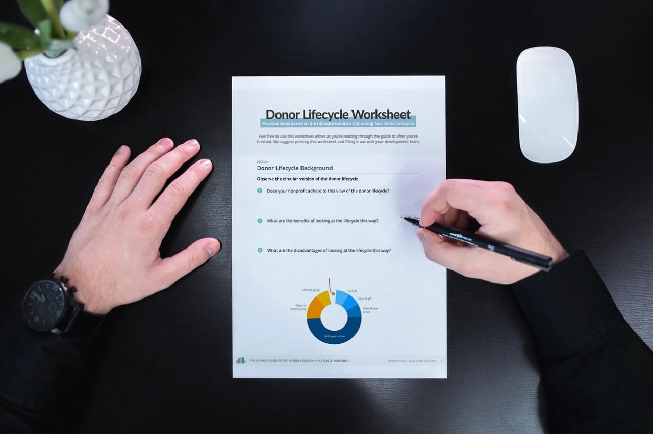 Person writing on a Donor Lifecycle Worksheet with a pen on a black desk with a mouse and plant.