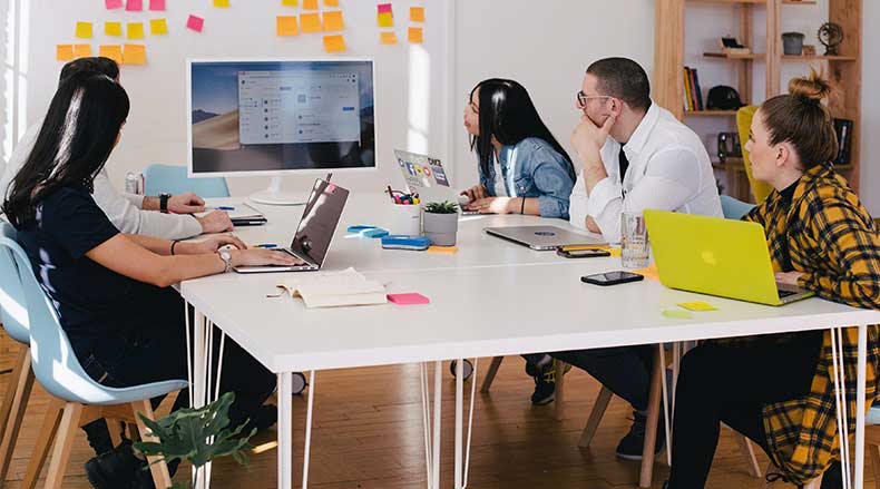 Five people seated around a white table looking at a large monitor in a bright office with post-it notes on wall.