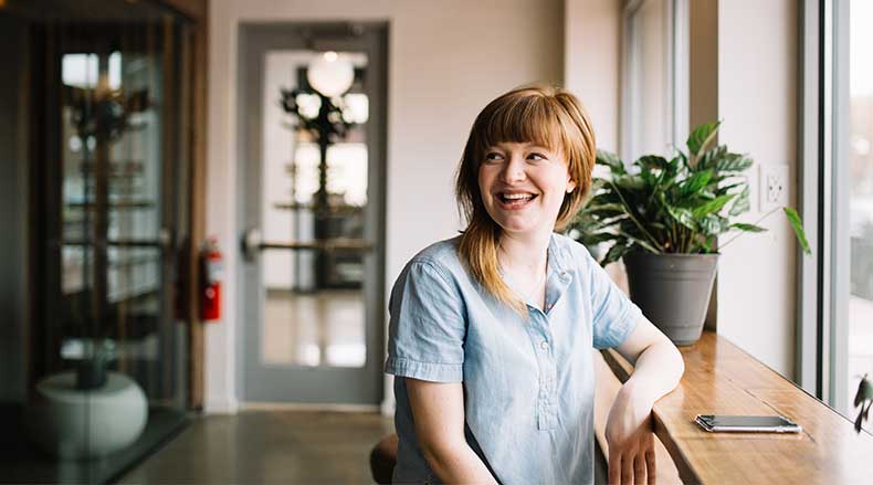 Smiling woman in blue shirt sitting at a wooden table by a window with a green plant and phone.