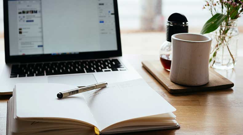 Open notebook with pen on a wooden desk, beside a laptop, coffee mug, and small flower vase.