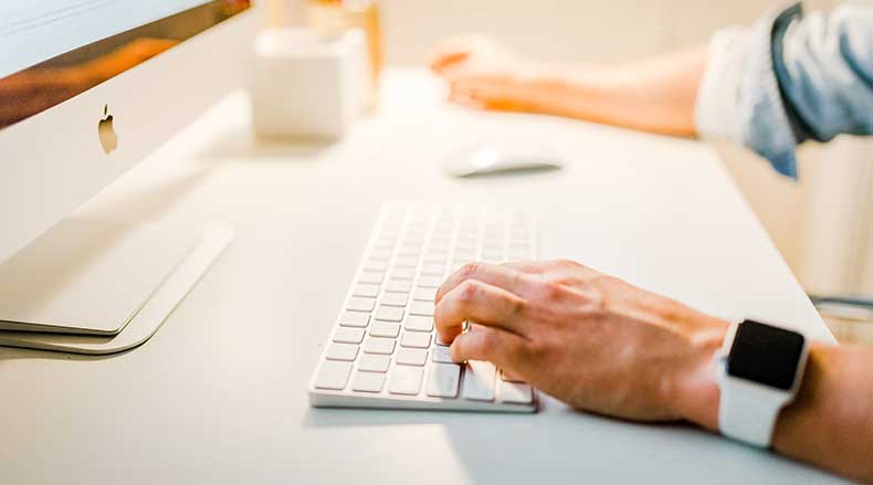 Person typing on a white wireless keyboard with an Apple computer and smartwatch visible.