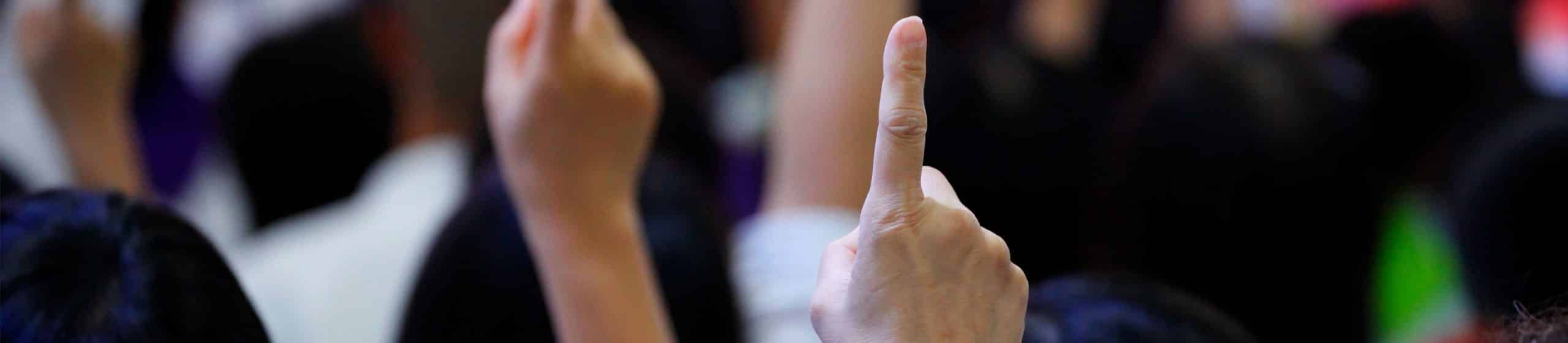 Raised hands with one hand's index finger pointing up among a blurred crowd.