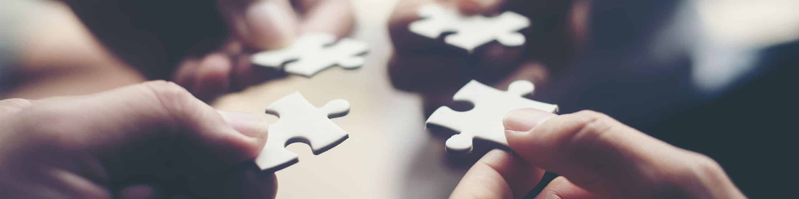 Four hands each holding a white puzzle piece close to fitting them together on a light surface.