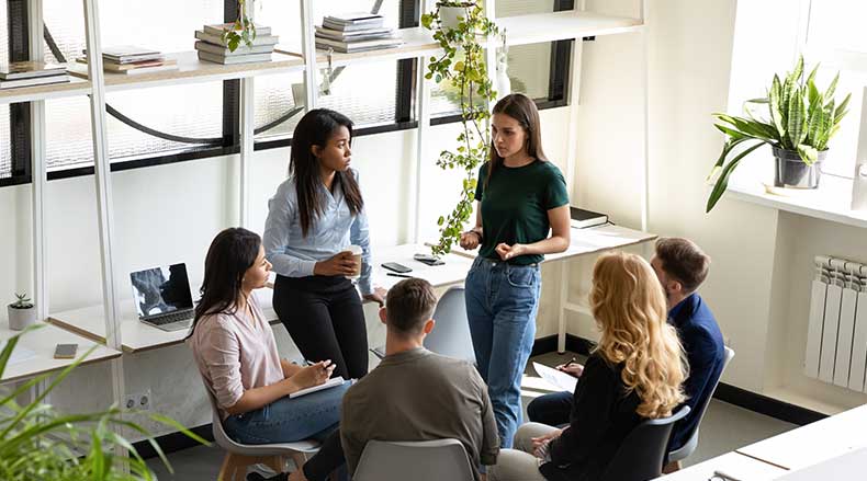 Group of six people having a discussion in a bright office space with plants and shelves.
