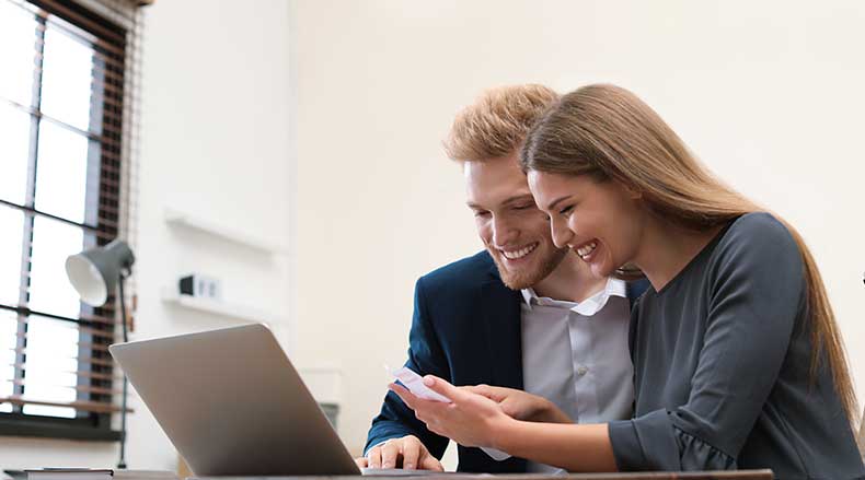 A man and woman smiling while looking at a smartphone together with a laptop on the table.
