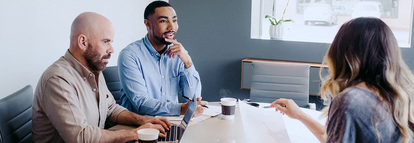 Three colleagues in a meeting room discussing documents and working on a laptop at a table.