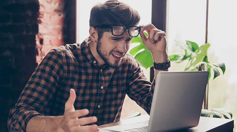 Young man in plaid shirt looking frustrated and lifting glasses while working on a laptop indoors.