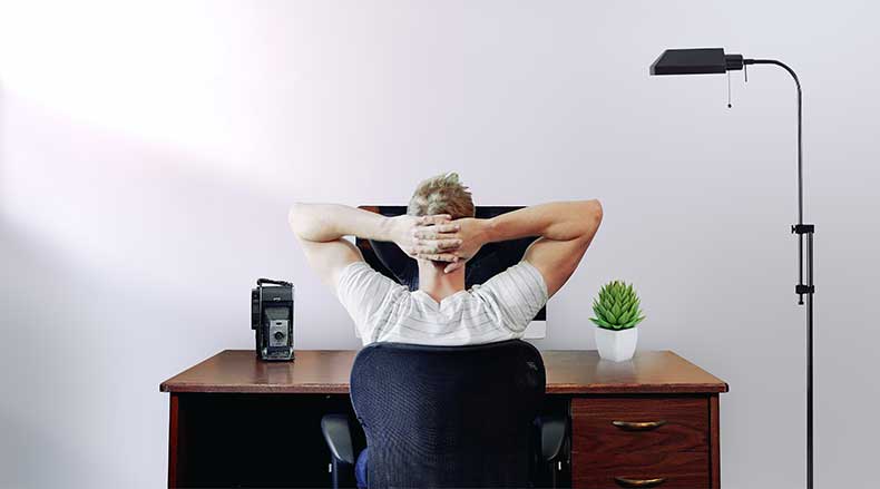Person sitting at desk with hands behind head, facing a computer screen, with a lamp and plant nearby.