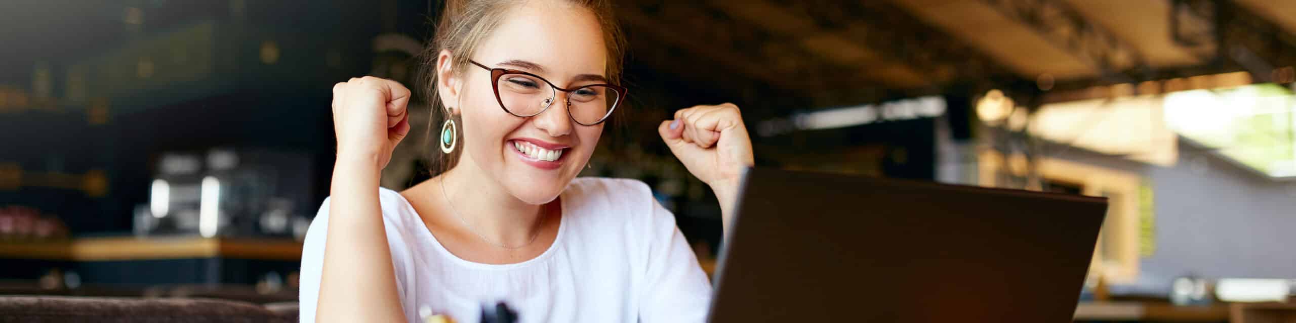 Smiling woman with glasses and earrings fist-pumping in excitement while looking at a laptop.