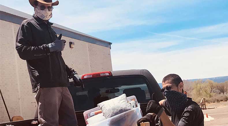 Two masked people loading household items from a pickup truck outdoors on a sunny day.