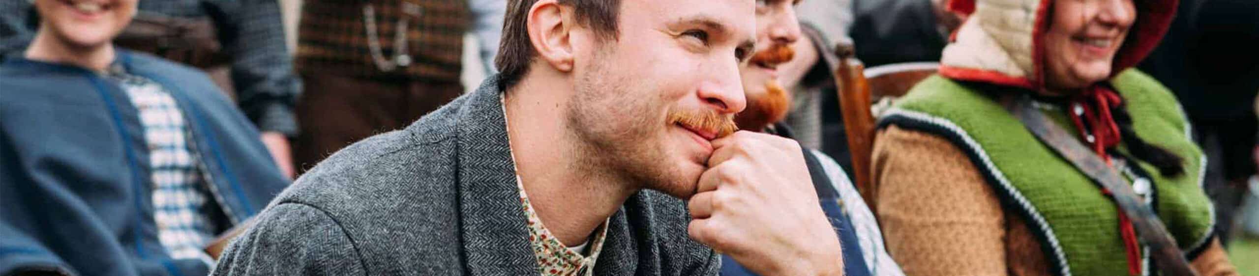A man with a mustache resting his chin on his fist, smiling, with others in background at an outdoor event.