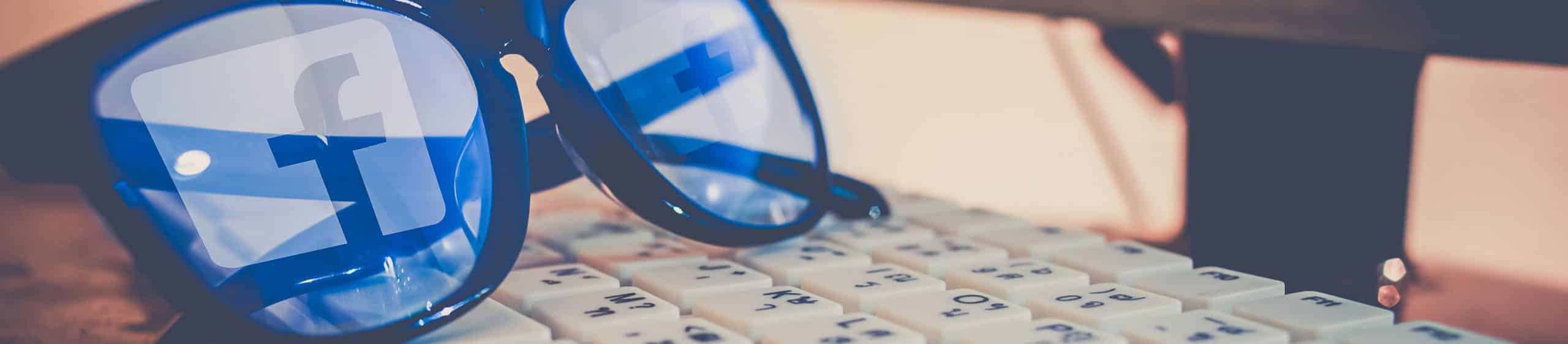 Close-up of glasses reflecting Facebook logos resting on a keyboard.
