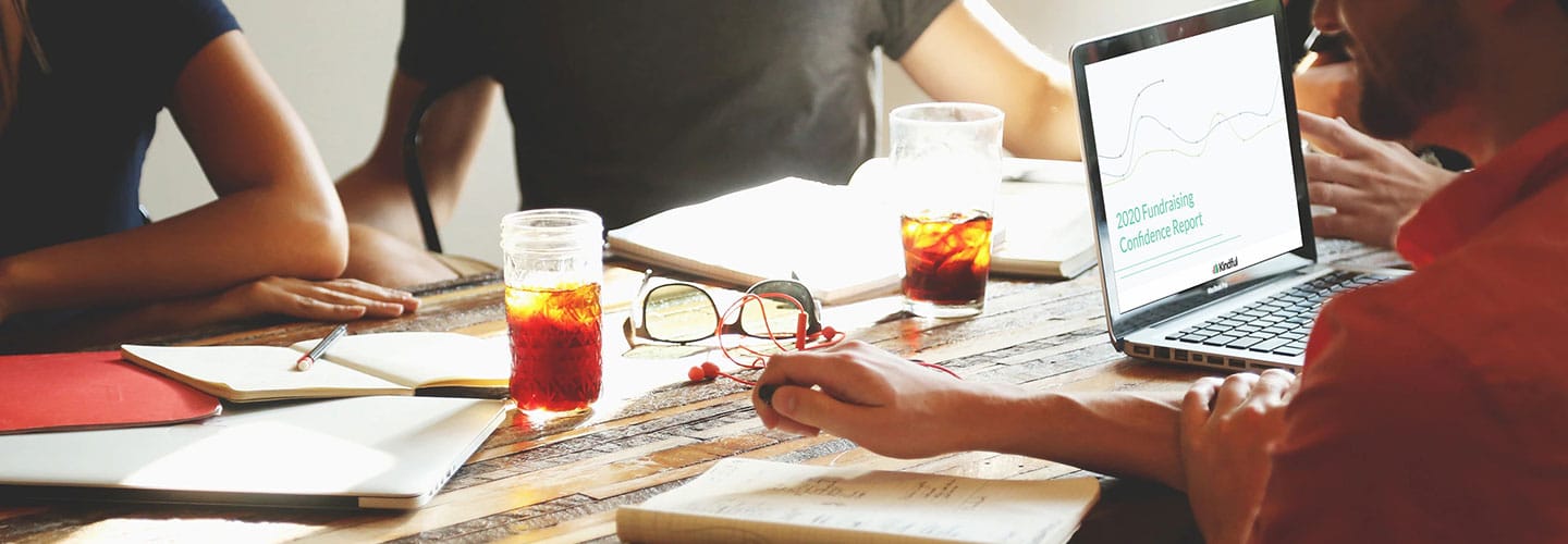 People at a wooden table with iced drinks, notebooks, glasses, and a laptop showing a fundraising report.