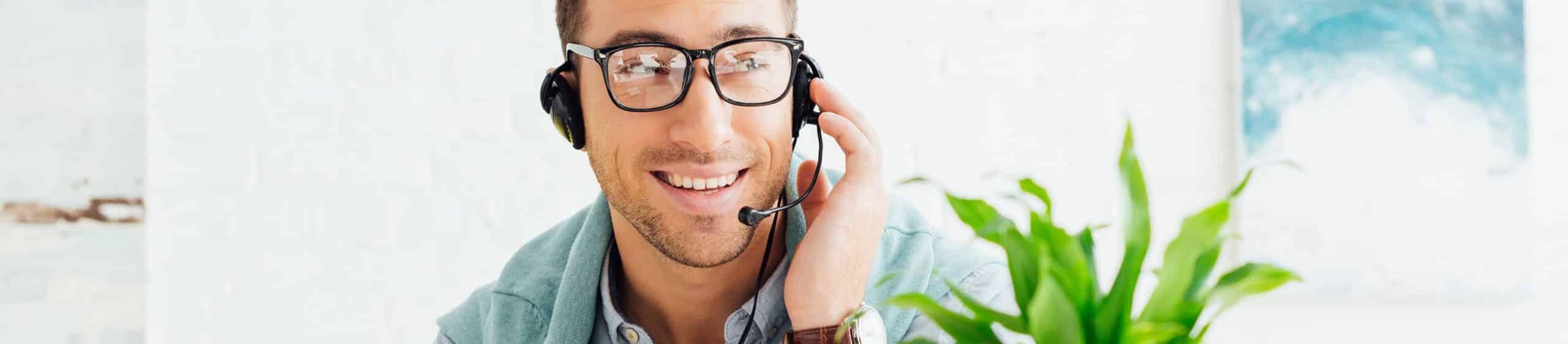 Smiling man wearing glasses and headset, holding headset microphone, with green plant nearby indoors.