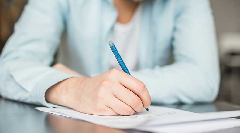Person in light blue shirt writing on white paper with a blue pen at a desk.