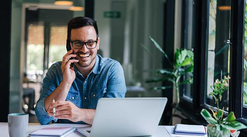 Man wearing glasses, smiling and talking on phone while working on a laptop in a bright room.