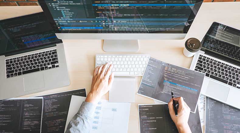 Person working on coding with multiple screens and printed code sheets on desk with coffee cup.