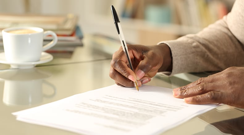 Close-up of a person signing documents with a pen at a glass table with a cup of coffee nearby.