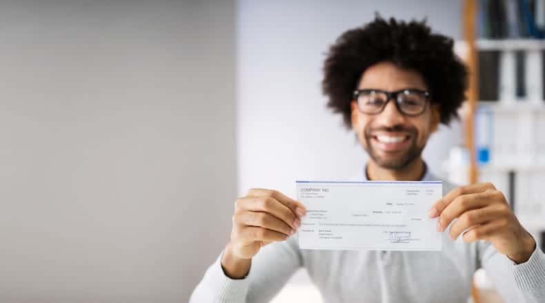 Smiling man with glasses holding a blank check towards the camera in an office setting.