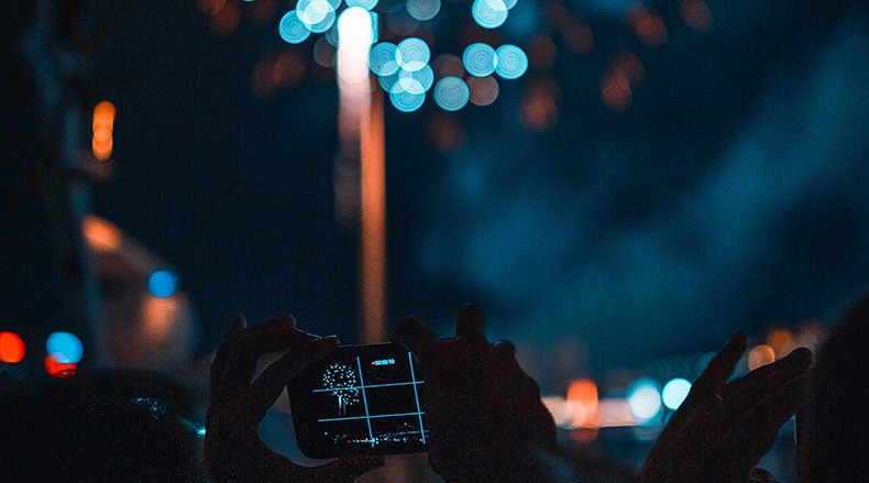 Person holds smartphone capturing fireworks at night with colorful blurred city lights in background.