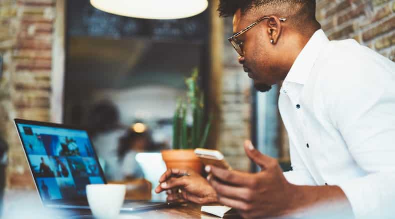 Man in white shirt using laptop and holding phone at a wooden table with cup and plant.