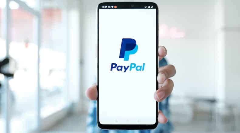Hand holding smartphone showing the PayPal logo on the screen in a blurred indoor setting.
