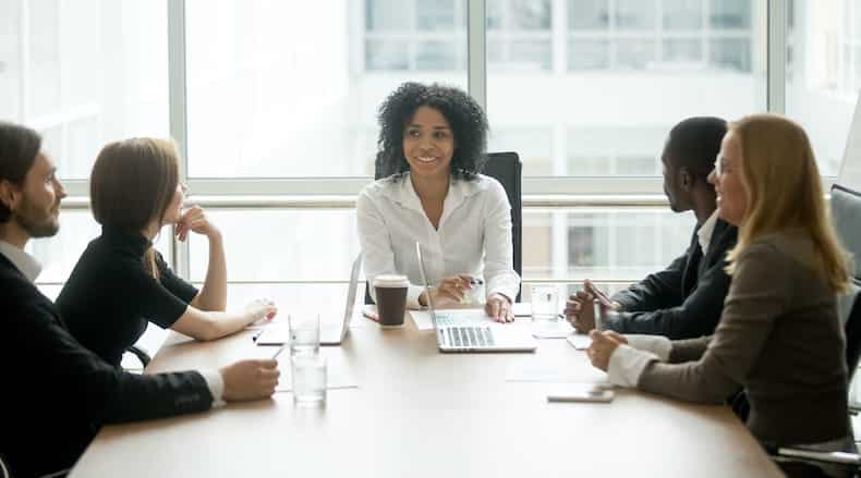 Smiling woman leading a meeting with four diverse colleagues around a conference table by a window.