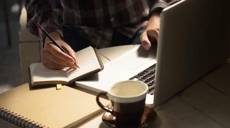 Person writing in a notebook while using a laptop, with a coffee cup on the table.