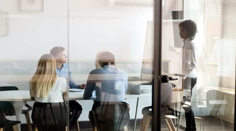 People meeting in a glass-walled office with one person standing and presenting at a whiteboard.