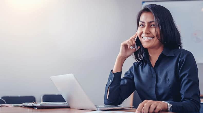 Smiling woman in dark shirt talking on phone at desk with open laptop and notebook.