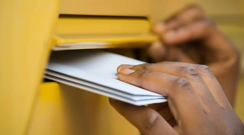 Person's hands inserting white envelopes into a yellow mailbox slot.