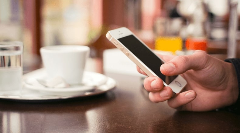 Hand holding smartphone over a table with a white coffee cup and glass of water in soft focus.