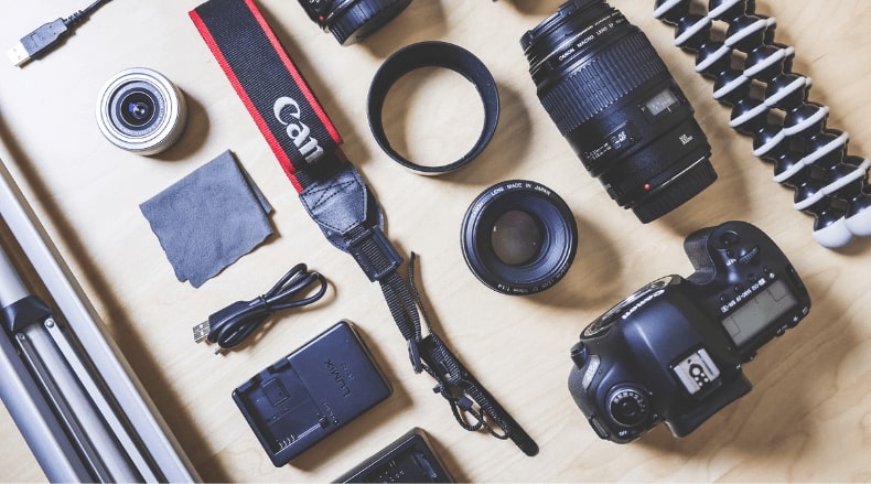 Camera gear including lenses, Canon strap, charger, USB cable, and tripod on a wooden surface.