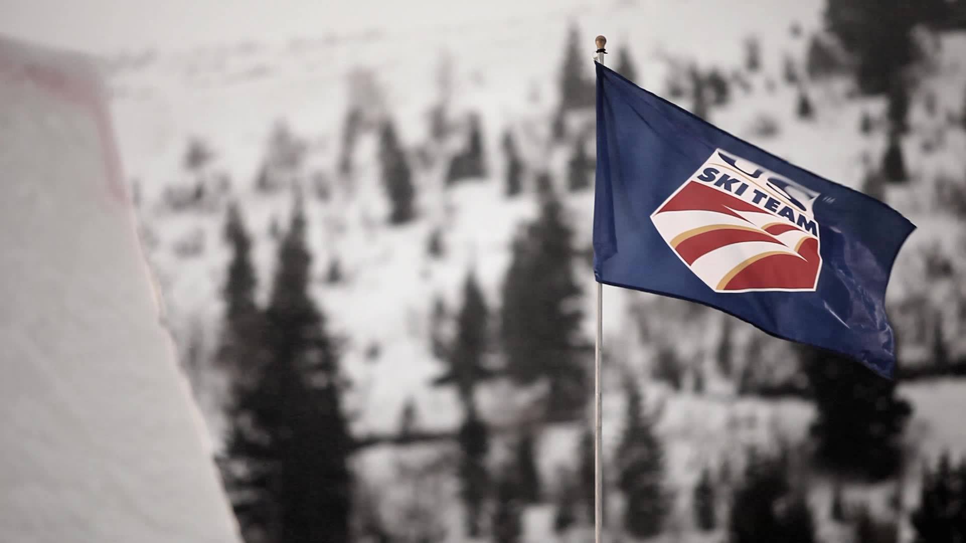 Blue flag with USA Ski Team logo flying on a pole against snowy mountain and trees background.