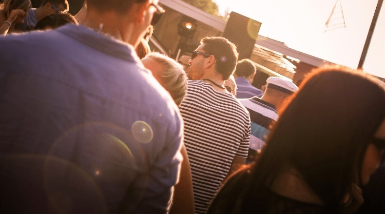 Crowd of people standing outdoors in warm sunlight, some wearing glasses and striped shirts.