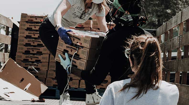 Three volunteers wearing masks and gloves unload boxes of food from a truck for donation.