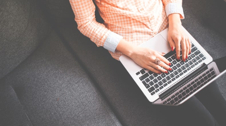 Person in a checkered shirt typing on a laptop while sitting on a dark gray couch.