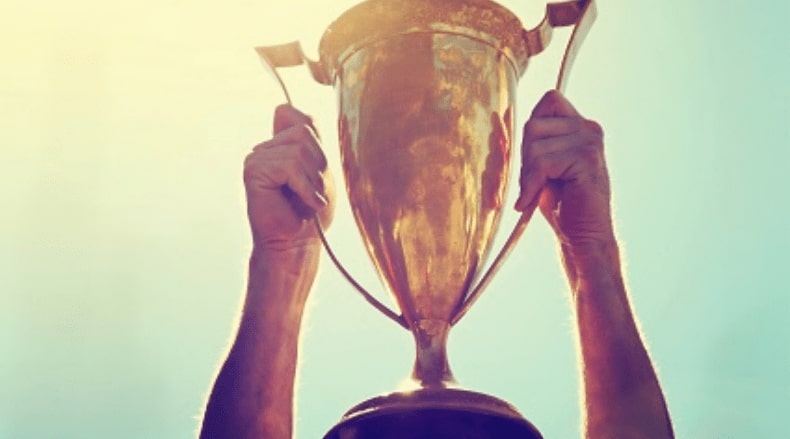 Two hands holding up a large golden trophy against a clear blue sky with sunlight.