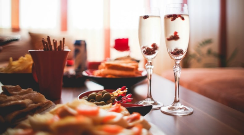 Two champagne flutes with floating berries on a table with appetizers and snacks in a cozy setting.