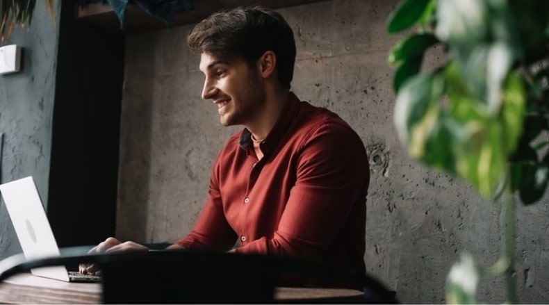 Smiling man wearing a red shirt working on a laptop at a desk near a concrete wall and plant.