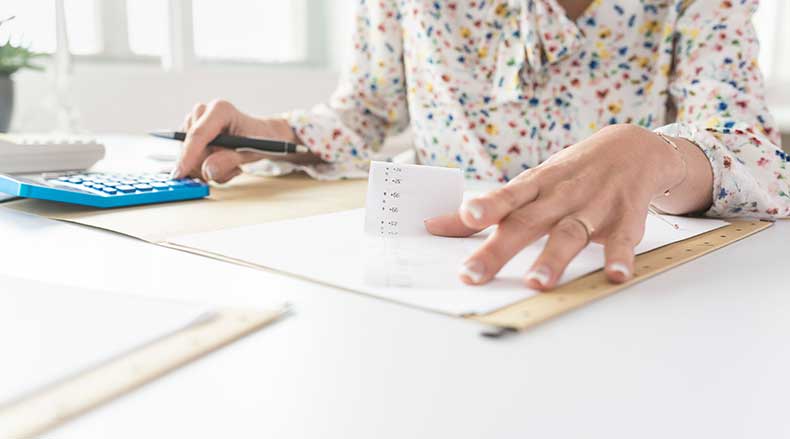 Person using calculator and pen to review a printed receipt with a ruler on desk.