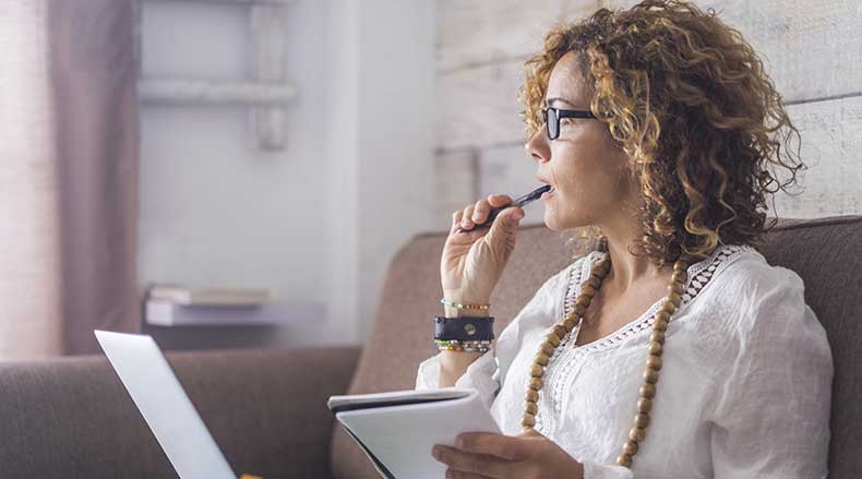 Woman with curly hair and glasses sitting on couch, holding notebook and pen near mouth, thinking.